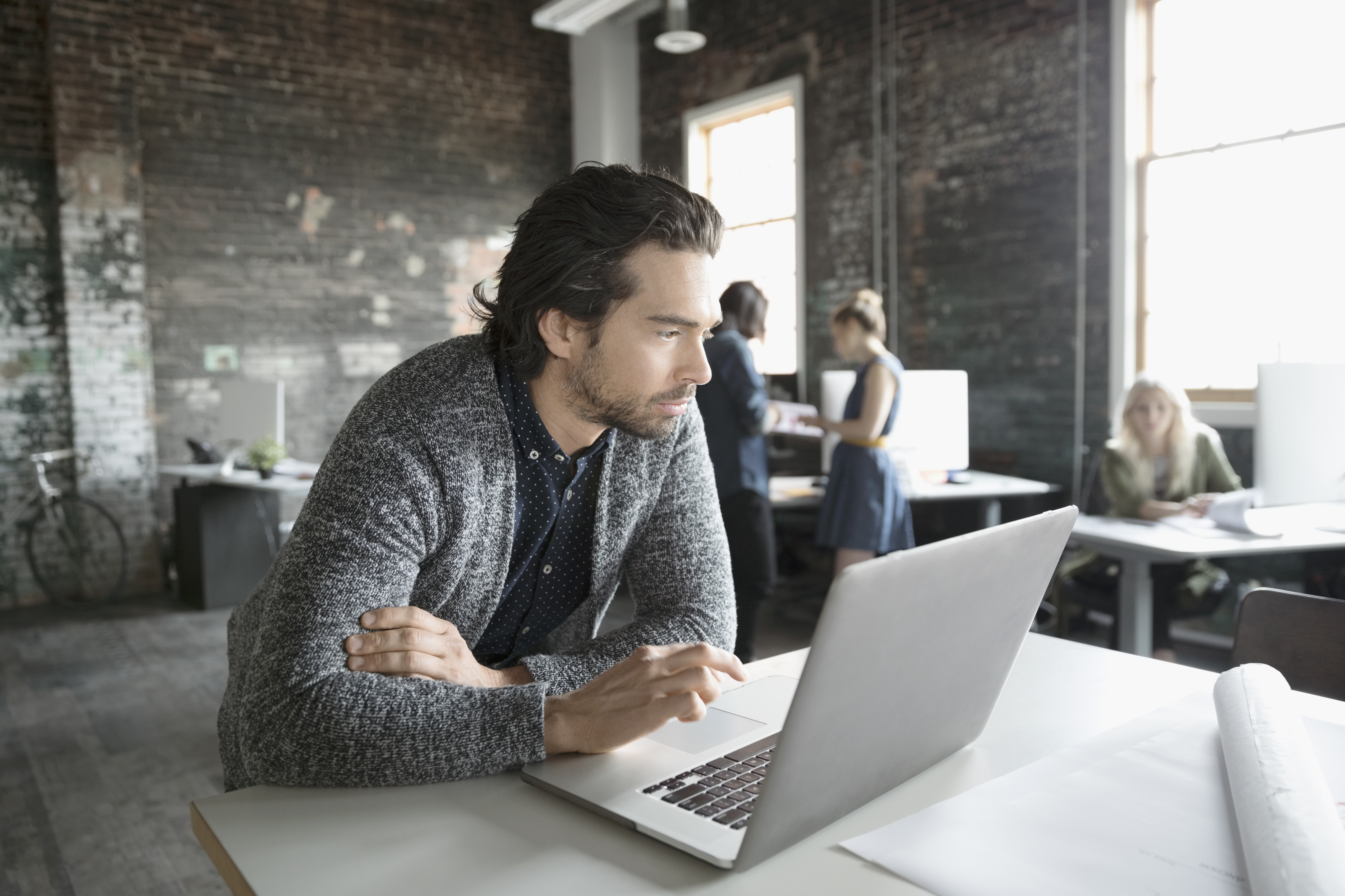 Man wearing a grey sweater working at a laptop in an office.