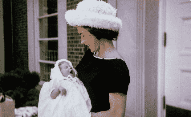 Woman in white hat and black dress holding a baby in white gown.