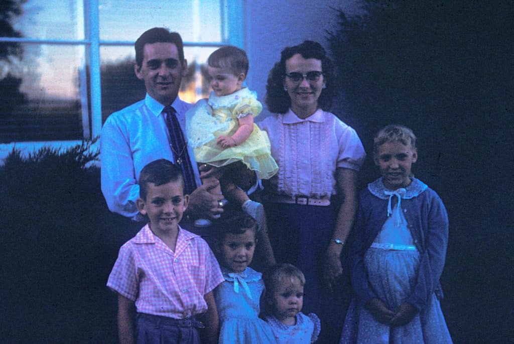 A photograph of a family standing together in front of a house in the 1950s whose color has become distorted over time, taking on a bluish tint
